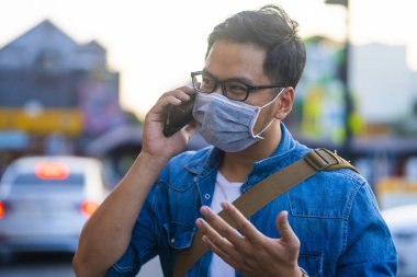 Sokakta telefon kullanırken ameliyat maskesi takan bir adam. Mikropları, zehirli dumanları ve tozu önlemek için koruyucu maske takan genç bir adamın portresi. Bakteriyel enfeksiyonun önlenmesi Corona virüsü veya Covid 19 sokaklarda ve bahçelerde.