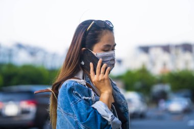 Sokakta cep telefonu kullanırken ameliyat maskesi takan genç bir kadın. Mikropları, zehirli dumanları ve tozu önlemek için koruyucu maske takan genç bir kadının portresi. Bakteriyel enfeksiyonun önlenmesi Corona virüsü veya Covid 19.