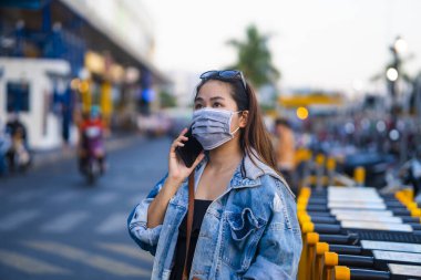 Sokakta cep telefonu kullanırken ameliyat maskesi takan genç bir kadın. Mikropları, zehirli dumanları ve tozu önlemek için koruyucu maske takan genç bir kadının portresi. Bakteriyel enfeksiyonun önlenmesi Corona virüsü veya Covid 19.