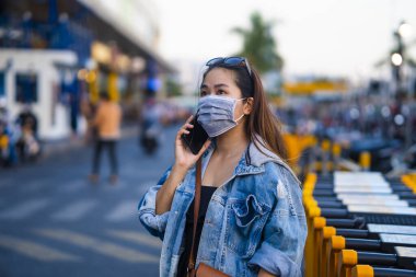 Sokakta cep telefonu kullanırken ameliyat maskesi takan genç bir kadın. Mikropları, zehirli dumanları ve tozu önlemek için koruyucu maske takan genç bir kadının portresi. Bakteriyel enfeksiyonun önlenmesi Corona virüsü veya Covid 19.