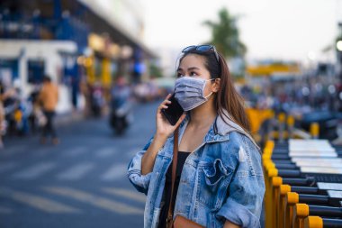 Sokakta cep telefonu kullanırken ameliyat maskesi takan genç bir kadın. Mikropları, zehirli dumanları ve tozu önlemek için koruyucu maske takan genç bir kadının portresi. Bakteriyel enfeksiyonun önlenmesi Corona virüsü veya Covid 19.