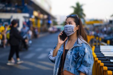 Sokakta cep telefonu kullanırken ameliyat maskesi takan genç bir kadın. Mikropları, zehirli dumanları ve tozu önlemek için koruyucu maske takan genç bir kadının portresi. Bakteriyel enfeksiyonun önlenmesi Corona virüsü veya Covid 19.