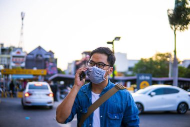 Sokakta telefon kullanırken ameliyat maskesi takan bir adam. Mikropları, zehirli dumanları ve tozu önlemek için koruyucu maske takan genç bir adamın portresi. Bakteriyel enfeksiyonun önlenmesi Corona virüsü veya Covid 19 sokaklarda ve bahçelerde.
