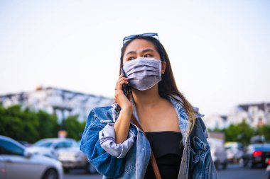Sokakta cep telefonu kullanırken ameliyat maskesi takan genç bir kadın. Mikropları, zehirli dumanları ve tozu önlemek için koruyucu maske takan genç bir kadının portresi. Bakteriyel enfeksiyonun önlenmesi Corona virüsü veya Covid 19.