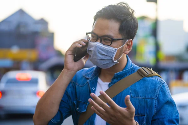 Man wearing surgical mask on street while using phone. Portrait of young man wearing a protective mask to prevent germs, toxic fumes, and dust. Prevention of bacterial infection Corona virus or Covid 19 in the air around the streets and gardens.