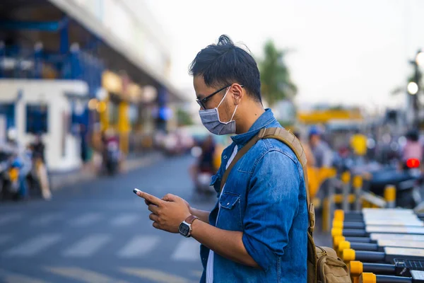 Man wearing surgical mask on street while using phone. Portrait of young man wearing a protective mask to prevent germs, toxic fumes, and dust. Prevention of bacterial infection Corona virus or Covid 19 in the air around the streets and gardens.