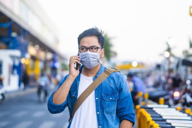 Sokakta telefon kullanırken ameliyat maskesi takan bir adam. Mikropları, zehirli dumanları ve tozu önlemek için koruyucu maske takan genç bir adamın portresi. Bakteriyel enfeksiyonun önlenmesi Corona virüsü veya Covid 19 sokaklarda ve bahçelerde.