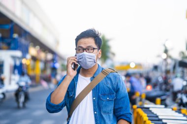 Sokakta telefon kullanırken ameliyat maskesi takan bir adam. Mikropları, zehirli dumanları ve tozu önlemek için koruyucu maske takan genç bir adamın portresi. Bakteriyel enfeksiyonun önlenmesi Corona virüsü veya Covid 19 sokaklarda ve bahçelerde.