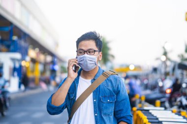 Sokakta telefon kullanırken ameliyat maskesi takan bir adam. Mikropları, zehirli dumanları ve tozu önlemek için koruyucu maske takan genç bir adamın portresi. Bakteriyel enfeksiyonun önlenmesi Corona virüsü veya Covid 19 sokaklarda ve bahçelerde.