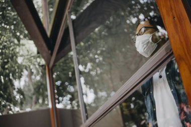 Üzgün adam, izole edilmiş genç adam, koruyucu steril tıbbi maskeyle evde kalıp pencereye bakıyor, sıkılmış çocuk. Karantina, enfeksiyonu önle. 