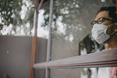 Üzgün adam, izole edilmiş genç adam, koruyucu steril tıbbi maskeyle evde kalıp pencereye bakıyor, sıkılmış çocuk. Karantina, enfeksiyonu önle. 