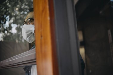 Üzgün adam, izole edilmiş genç adam, koruyucu steril tıbbi maskeyle evde kalıp pencereye bakıyor, sıkılmış çocuk. Karantina, enfeksiyonu önle. 