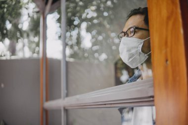 Üzgün adam, izole edilmiş genç adam, koruyucu steril tıbbi maskeyle evde kalıp pencereye bakıyor, sıkılmış çocuk. Karantina, enfeksiyonu önle. 