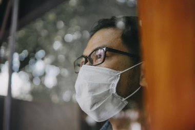 Üzgün adam, izole edilmiş genç adam, koruyucu steril tıbbi maskeyle evde kalıp pencereye bakıyor, sıkılmış çocuk. Karantina, enfeksiyonu önle. 