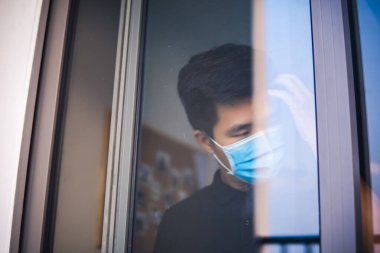 Kendini karantinaya al. Pencereden dışarı bakan tıbbi maskeli üzgün bir genç adam. Kendini izole eden tıbbi maskeli adam bir evin penceresinden sokağa bakıyor.