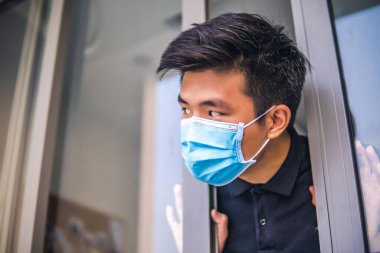 Tıbbi maskeli genç Asyalı adam evin kapısını açıyor. Coronavirus konsepti. Solunum koruması. Evde karantina.
