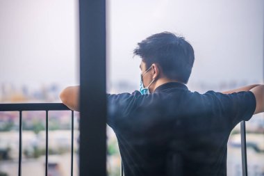 Kendini karantinaya al. Pencereden dışarı bakan tıbbi maskeli üzgün bir genç adam. Kendini izole eden tıbbi maskeli adam bir evin penceresinden sokağa bakıyor.