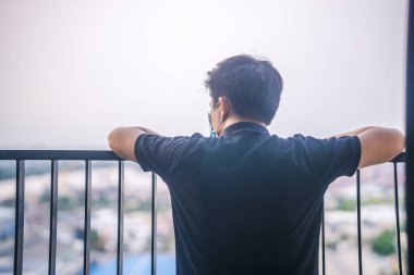 Kendini karantinaya al. Pencereden dışarı bakan tıbbi maskeli üzgün bir genç adam. Kendini izole eden tıbbi maskeli adam bir evin penceresinden sokağa bakıyor.