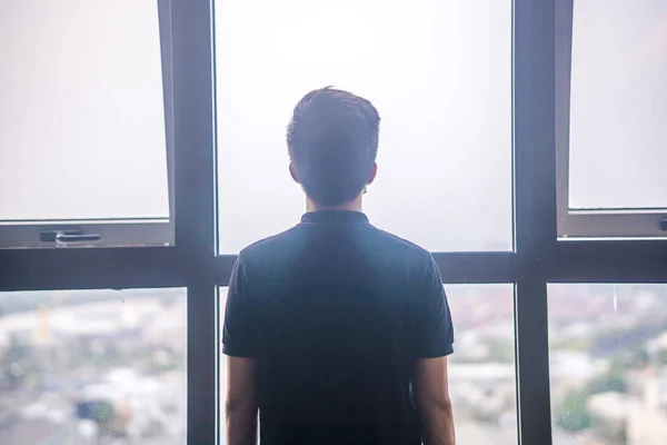 Quarantine self-isolation. Sad young man in a medical mask who looks out the window through the window. Infected man in medical mask on self-isolation looks at the street through the window of a house