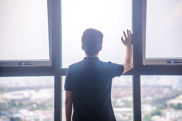 Quarantine self-isolation. Sad young man in a medical mask who looks out the window through the window. Infected man in medical mask on self-isolation looks at the street through the window of a house