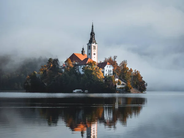 Fog over the water of Bled Lake in front of the church on a small island and in the background Bled Castle, built on high cliffs, with reflection in the lake on a beautiful autumn morning in Slovenia