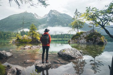 Dağlara bakan kırmızı ceketli, şapkalı ve sırt çantalı bir adamın manzarası, Hintersee Gölü, Berchtesgaden, Almanya
