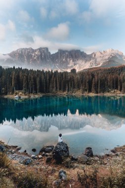Dolomitler, İtalya 'da, Karersee Gölü' nde mavi gökyüzüne karşı mutlu sarışın kızın arka görüntüsü. Çiçekli elbisesi ve şapkası olan kaygısız kadın dağlarla çevrili göl manzarasının tadını çıkarıyor.