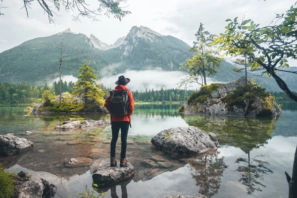 Dağlara bakan kırmızı ceketli, şapkalı ve sırt çantalı bir adamın manzarası, Hintersee Gölü, Berchtesgaden, Almanya