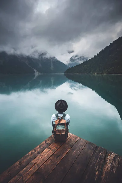 身着斜纹棉布夹克 头戴黑色帽子 背着棕色背包的旅行者们 在闷热的日出时分 坐在湖上的木桥上 奥地利蒂罗尔普朗西湖的旅行和积极生活概念 — 图库照片