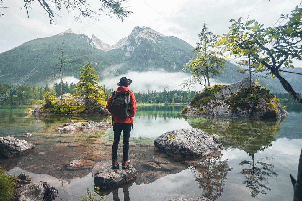 Scenic view of a man wearing a red jacket, a hat and a backpack looking at mountains, Lake Hintersee, Berchtesgaden, Germany