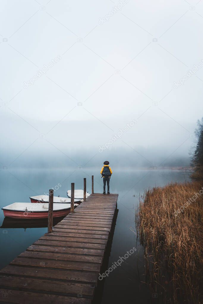 Backpacker standing on pier on a moody sunrise at Lake Sfanta Ana with anchored boats against foggy mountains