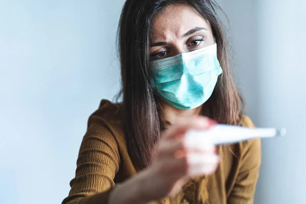 Cerrahi maske takan hasta genç bir kadın ve elinde 38 dereceden fazla ateşi olduğunu gösteren dijital bir termometre var. Salgın sırasında ateşi ve hastalığı olan hasta ve endişeli bir kadın. Karantina, kendini izole etme ve koronavirüs konsepti. Baş ağrısı. Grip.