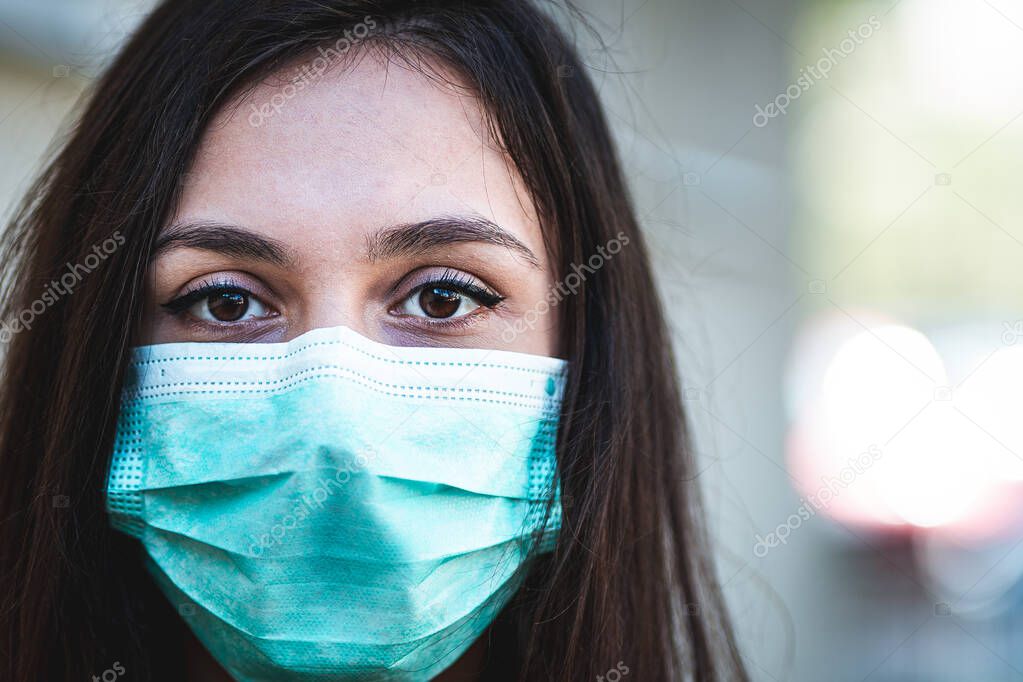 Close up portrait of Latino young woman wearing a protective surgical face mask and looking at camera, she is looking optimistic about epidemic end
