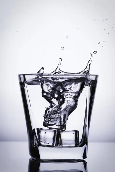 Παγάκια εκτινάξεις σε ποτήρι, παγάκι έπεσε σε ένα ποτήρι νερό, φρέσκο, κρύο νερό, απομονώνονται σε λευκό, μπλε, ασημί, μπλε φόντο. — Φωτογραφία Αρχείου