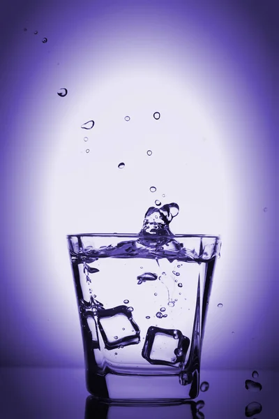 Cubos de hielo salpicando en vidrio, cubo de hielo caído en un vaso de agua, agua dulce y fría, aislado en violeta, fondo azul violeta . —  Fotos de Stock