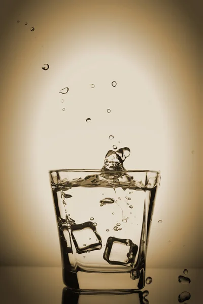 Παγάκια εκτινάξεις σε ποτήρι, παγάκι έπεσε σε ένα ποτήρι νερό, φρέσκο, κρύο νερό, απομονώνονται σε μπρούτζινο, κίτρινο φόντο. — Φωτογραφία Αρχείου