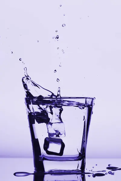 Ijsblokjes spatten in glas, daalde ijsblokje in een glas water, vers, koud water, geïsoleerd op violet blauw, violet achtergrond. — Stockfoto