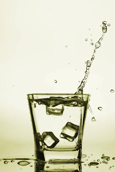 Παγάκια εκτινάξεις σε ποτήρι, παγάκι έπεσε σε ένα ποτήρι νερό, φρέσκο, κρύο νερό, απομονώνονται σε φόντο χρυσό, κίτρινο, χάλκινο. — Φωτογραφία Αρχείου