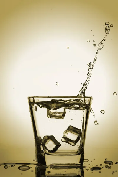 Παγάκια εκτινάξεις σε ποτήρι, παγάκι έπεσε σε ένα ποτήρι νερό, φρέσκο, κρύο νερό, απομονώνονται σε χάλκινο, χρυσό, κίτρινο φόντο. — Φωτογραφία Αρχείου