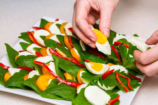 Kadın eller ıspanak yumurta kırmızı biber ile yeşil salata yapmak, beyaz plaka üzerinde domates ve peynir, kelebekler ile gri masa örtüsü üzerinde, sağlıklı gıda, sağlıklı yaşam. — Stok fotoğraf