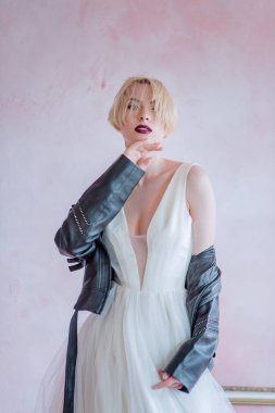 Güzel gelin kamerada poz veriyor. Kısa saçlı, gelinlikli ve deri ceketli bir kız..