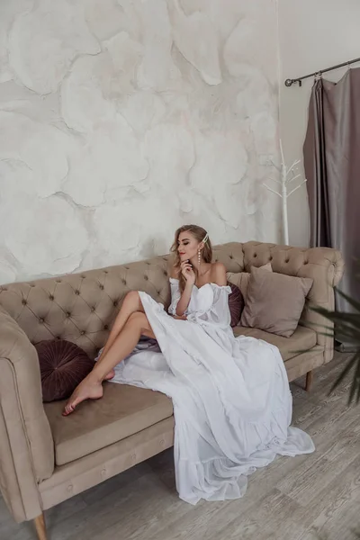 ファッション花嫁でファッショナブルな白いドレスとともに彼女の髪と長いイヤリング現代的なソファに座って — ストック写真