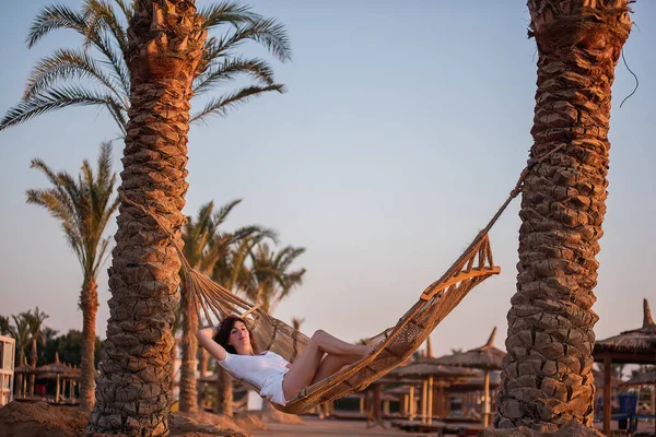 A beautiful girl on a hammock holiday between palm trees and the sea. I dream of a journey after quarantine. Remote work.