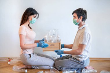 Tıbbi maskeli genç bir kadın ve tek kullanımlık mavi eldivenli genç bir adam karşı karşıya oturmuş, boş plastik şişelerle dolu ahşap bir kutu tutuyorlar - eko-plastik geri dönüşüm.