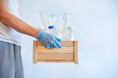 Mavi tek kullanımlık eldivenlerle el ele tutuşan kişi içinde çeşitli boş eko-plastik şişelerin bulunduğu ahşap bir kutu. Plastik konsepti düzenliyor.