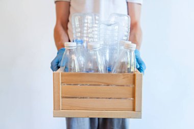 Mavi tek kullanımlık eldivenlerle el ele tutuşan kişi içinde çeşitli boş eko-plastik şişelerin bulunduğu ahşap bir kutu. Plastik konsepti düzenliyor.