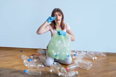 Genç kadın üzgün ve ailesinin etrafında bir sürü plastik olduğu için ağlıyor. Mavi eldivenli bir kız, çöpü şeffaf bir torbaya koymak için boş plastik kutular topluyor..