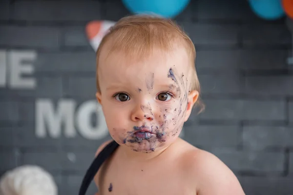 The first birthday in life is a large portrait of a satisfied little boy, the face is stained with the remains of the cake, the baby has tasted the sweet cake for the first time, on the background of festive colorful balls.