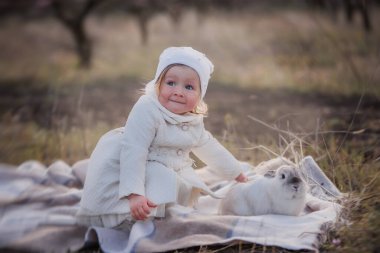 Küçük kız gri bir tavşanı okşuyor. Ekose bir ceket ve beyaz şapkayla oturan, evcil bir hayvanla piknik yapan, doğada tavşan gezdiren küçük bir sarışın..