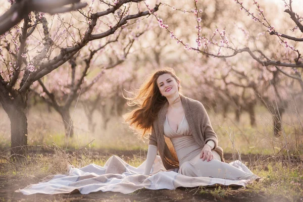 赤い長い髪の美しい女の子は 花の庭で遊んだ上に座っています 彼女の髪は風の中で発展し 彼女は笑顔で ベージュのドレスと茶色のニットカーディガンを着ています 家族でピクニック — ストック写真